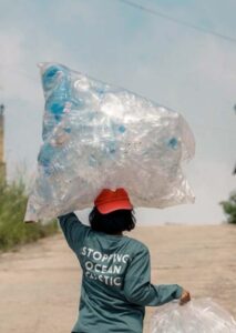 Eine Person trägt einen riesigen, transparenten Sack voller gesammelter leerer Plastikflaschen auf dem Kopf. Auf dem Tshirt der Person steht der Schriftzug"STOPPING OCEAN PLASTIC"