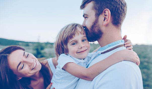 Ein lachender Vater umarmt sein lächelndes Kind, während die Mutter im Hintergrund ebenfalls lächelt und sie anschaut